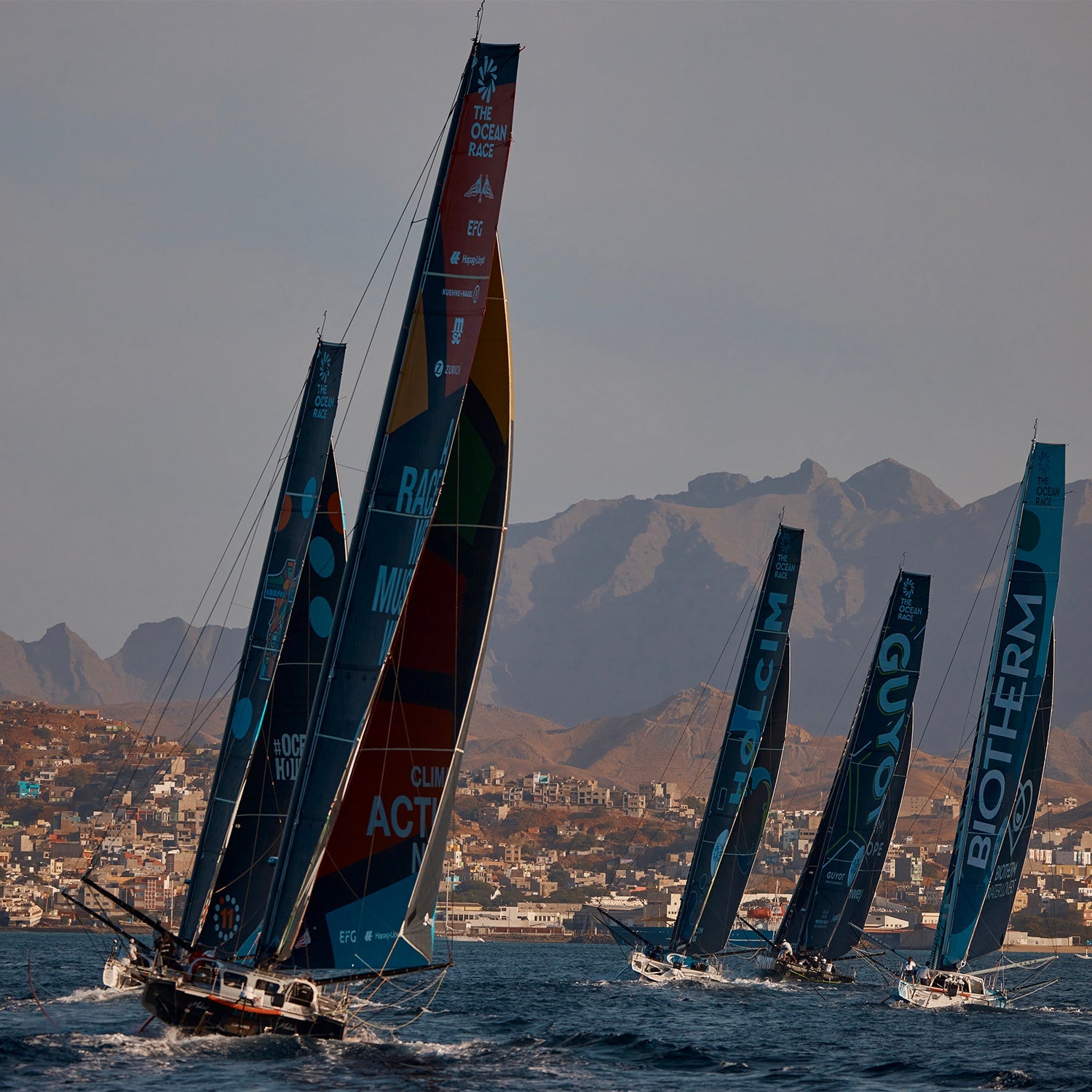 Five ships sailing at sea, with the town of Mindelo in the background, as they set off on the Ocean Race second leg 2023.//© GUYOT environment - Team Europe | Charles Drapeau