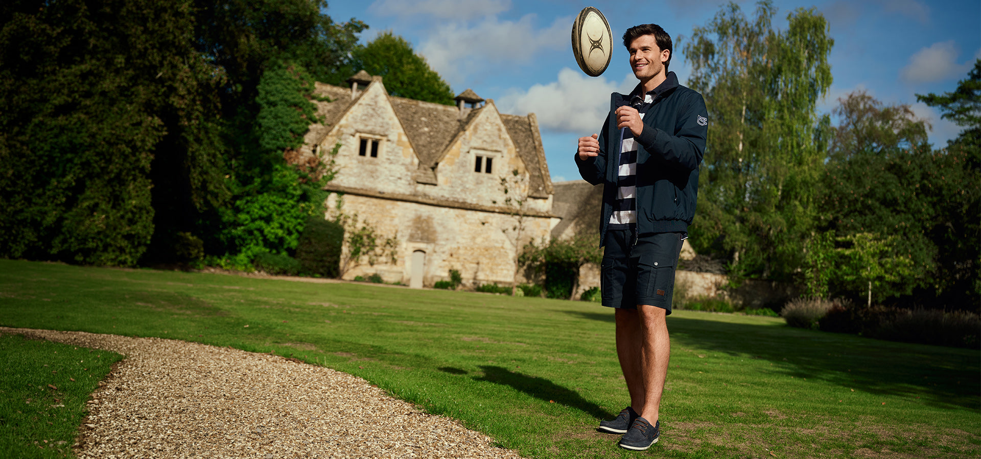Man holding a rugby ball in a scenic outdoor setting with a building and trees in the background