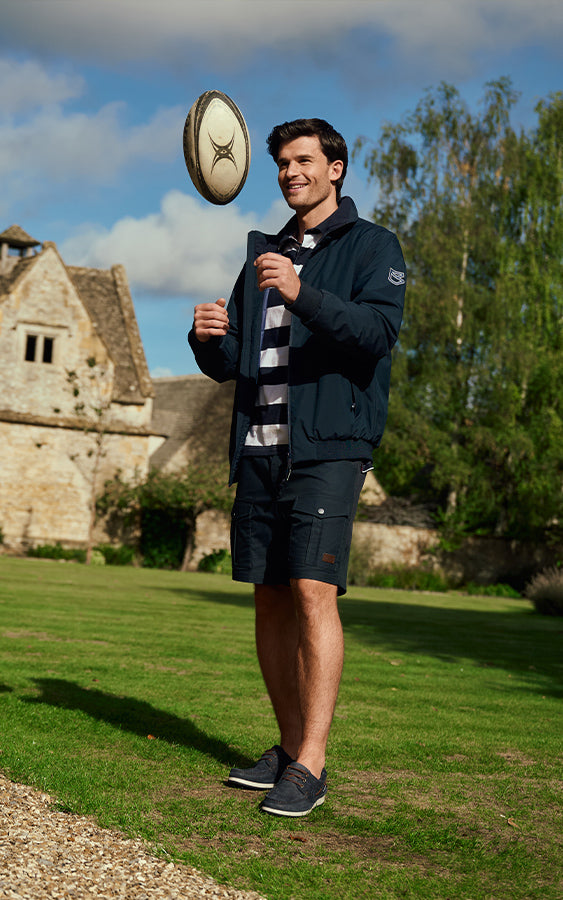 Man holding a rugby ball outdoors with a building and trees in the background