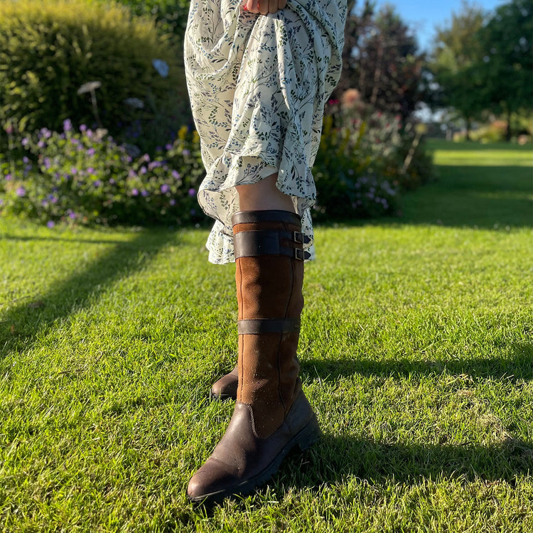 Square image of the Dubarry Longford Leather Waterproof and Breathable Country Boot being worn with a dress on a well-maintained lawn with some shrubs in the background.//© Bonnie Baker